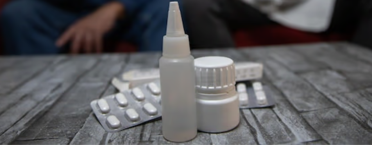 Uma mesa com medicamentos enquanto um médico conversa com seu paciente ao fundo.