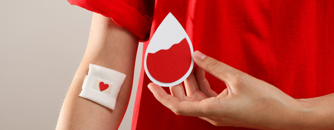 Pessoa com camisa vermelha e curativo adesivo em formato de coração no braço direito, segurando desenho de gota de sangue.
