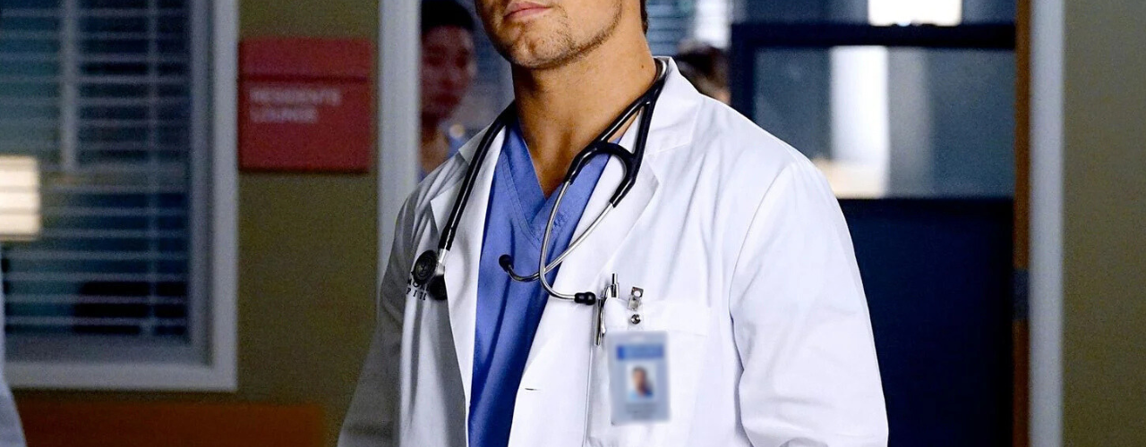 Médico jovem, vestindo jaleco branco e scrubs azuis, com um estetoscópio Littmann preto pendurado no pescoço.