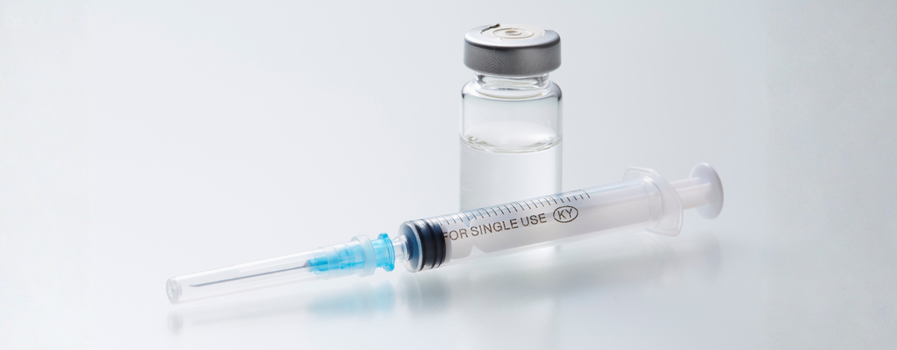 Frasco-ampola transparente ao lado de uma seringa esterilizada utilizada para aplicação de medicamentos injetáveis.