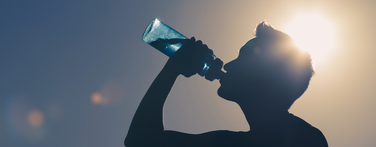 Homem bebendo água sob sol intenso, destacando a importância da hidratação para prevenir desidratação durante o verão.