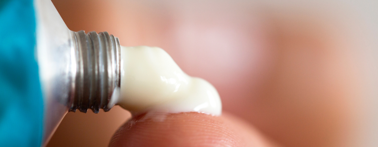 Bisnaga metálica aplicando creme branco sobre a pele, representando o uso de pomada anestésica antes de procedimento clínico.